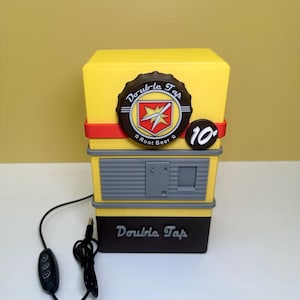 Könnte beinhalten: Eine gelb-schwarze Double Tap Root Beer-Maschinenförmige Lampe mit einem roten Streifen. Die Lampe hat ein braunes Emblem mit den Worten "Double Tap" und "Root Beer". Ein schwarzes Netzkabel und ein Steuerschalter sind angebracht.