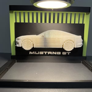 May include: A 3D-printed model of a Mustang GT car. The car is made of vertical, light-colored wooden slats, mounted on a black background with the text "MUSTANG GT". The display has a black frame and green accents.