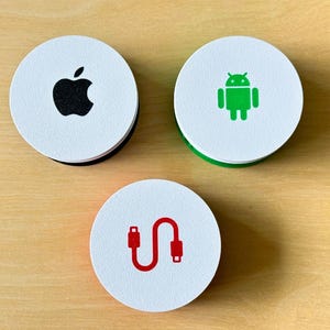 May include: Three round white coasters with black, green, and red graphics. One coaster features a black apple logo, another has a green Android figure, and the third displays a red cable icon. The coasters are on a light brown wooden surface.