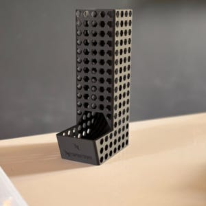 Op de afbeelding: Een zwarte Nespresso capsulehouder met een geometrisch ontwerp. De houder is rechthoekig met zeshoekige en cirkelvormige uitsparingen. De basis heeft het Nespresso-logo. De houder is gemaakt van matzwart materiaal en staat op een licht oppervlak.