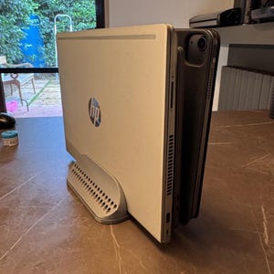 May include: A silver HP computer tower with the HP logo, holding a black tablet. The tower has a perforated base and a sleek, modern design. The tablet is positioned vertically, next to the tower.