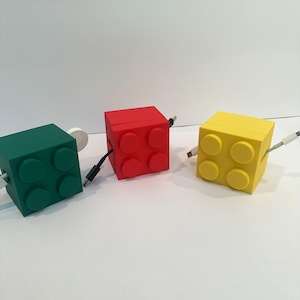 May include: Three colorful, cube-shaped cable organizers in green, red, and yellow. Each organizer has a design reminiscent of toy building blocks, with four raised circular studs on one side. Cables extend from the sides.
