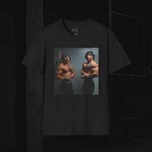 Puede incluir: Camiseta negra con una imagen cuadrada de dos hombres musculosos posando. Los hombres visten ropa oscura y tienen tatuajes. El fondo es un gimnasio. La camiseta tiene cuello redondo y mangas cortas.