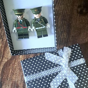 May include: Two toy soldier figures in olive green uniforms and hats are presented in a black and white polka dot gift box. The box is open, revealing the figures. A second gift box, also black with white polka dots, is tied with a white ribbon.