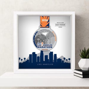 May include: Framed Los Angeles Marathon medal with a cityscape silhouette. The medal is silver and blue, with the text "2026" and "Los Angeles Marathon." Includes the text "March 6, 2026, DASH PARKER, 3:25:52, 26.2 FINISHER."
