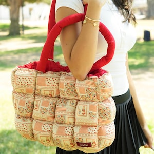 May include: A quilted tote bag with a red handle and a patchwork design. The bag features a repeating pattern of floral and geometric prints in cream, orange, and red. The word "Andan" is on a small tag.