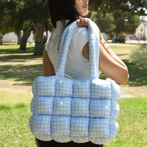 May include: A light blue and white gingham pattern tote bag with a puffy, quilted design. The bag has a rectangular shape with a padded handle. The bag is carried over the shoulder.