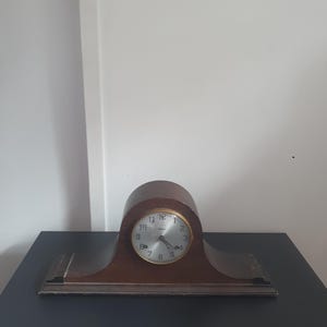 May include: A vintage wooden mantel clock with a curved top and base. The clock face is silver with black numerals and gold-colored trim. The clock is made of dark brown wood and sits on a black surface.