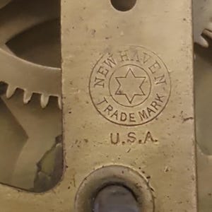 May include: Close-up of a brass clock mechanism with the text "NEW HAVEN TRADE MARK U.S.A." The clock part has a circular stamp with a star in the center. The gears and other components are visible.