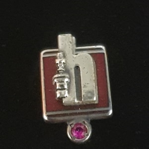 May include: A silver-toned pin with a red background and a stylized letter "H" design. A small, silver-toned object is on the left side of the "H". A small, round, pink gemstone is at the bottom of the pin.