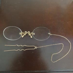 Puede incluir: Gafas de pinza antiguas de color dorado con lentes transparentes. Las gafas presentan un puente decorativo y una fina cadena. También se incluye una horquilla dorada a juego, probablemente para asegurar las gafas. El diseño recuerda a las gafas vintage.