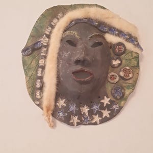 Könnte beinhalten: Eine Keramikmaske mit dunklem Gesicht, offenem Mund und Fellbesatz. Die Maske hat eine kreisförmige Form mit einem grünen, blattartigen Rand. Blaue und weiße Sterne verzieren den unteren Teil.