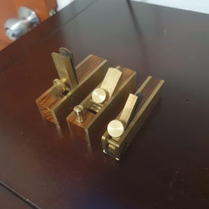 Puede incluir: Tres pequeños cepillos de mano con herrajes de latón y cuerpos de madera se muestran. Los cepillos son rectangulares, con cuchillas y perillas de ajuste. La madera tiene un color marrón rojizo.