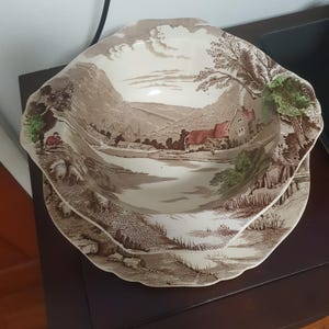 Może przedstawiać: Dwuczęściowy zestaw zabytkowych ceramicznych naczyń do serwowania. Naczynia przedstawiają brązową i kremową scenerię krajobrazu z domem, drzewami i wodą. Zestaw zawiera płytką miskę i talerz, oba z ząbkowanymi krawędziami.