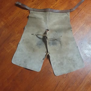 May include: A pair of worn, gray leather work chaps with a brown leather belt and straps. The chaps have a rectangular shape with rounded corners and are designed to protect the legs. Rivets secure the leather.