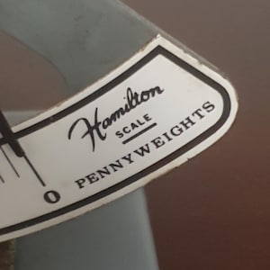 Puede incluir: Primer plano de una báscula Hamilton vintage. La etiqueta dice "Hamilton SCALE PENNY WEIGHTS" en escritura negra y letras de bloque sobre un fondo blanco con un borde negro. La báscula es de color azul claro.