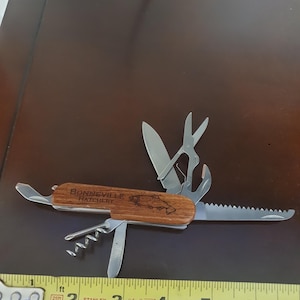 Puede incluir: Una multiherramienta con mango de madera, con un grabado de pez y el texto "BONNEVILLE HATCHERY". La herramienta incluye un cuchillo, tijeras y otros implementos. Una cinta métrica se coloca junto a la herramienta.