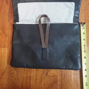May include: Black leather clutch with a brown strap and a white and black striped interior. The clutch is open, revealing the interior lining. A ruler is next to the clutch, measuring approximately 10 inches in length.