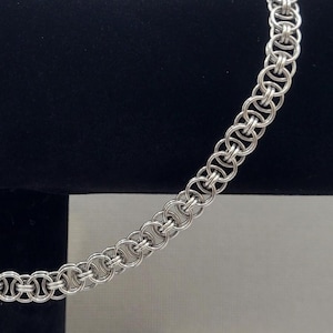 Peut inclure: Un collier en chaîne de couleur argentée avec un motif entrelacé détaillé. Le collier est présenté sur un présentoir noir sur fond neutre. La chaîne est composée de petits maillons circulaires.