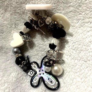 May include: A beaded bracelet with a variety of charms and beads in black, white, silver, and iridescent colors. The bracelet includes a heart-shaped charm, a flower-shaped charm, and a mix of round and faceted beads. The background is a soft, white fabric.