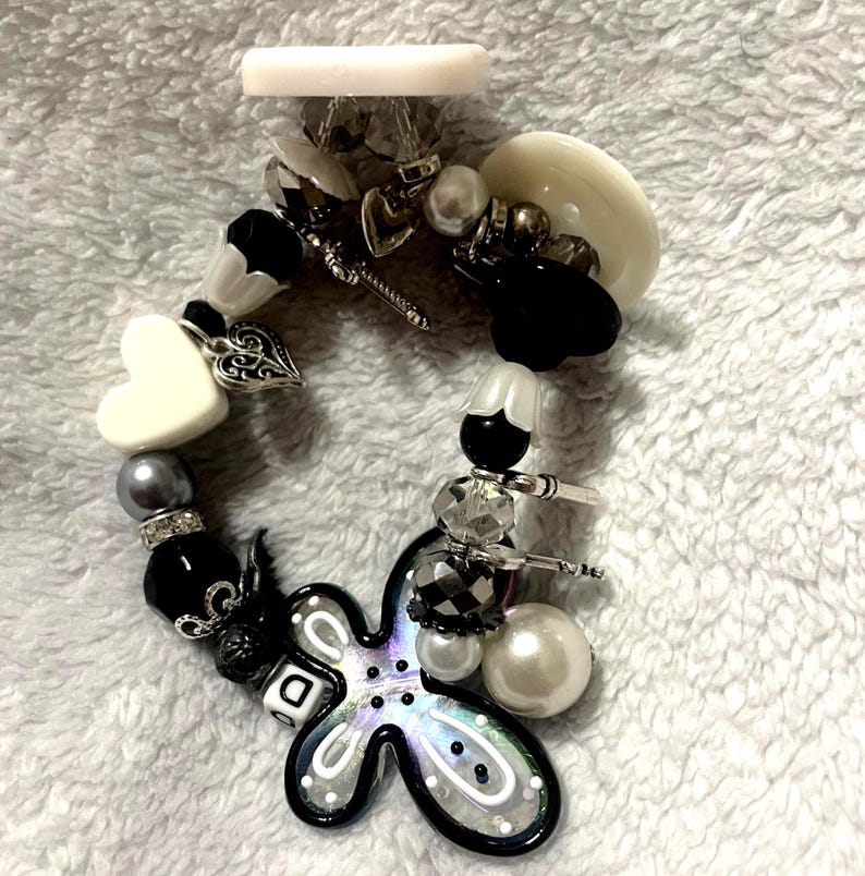 May include: A beaded bracelet with a variety of charms and beads in black, white, silver, and iridescent colors. The bracelet includes a heart-shaped charm, a flower-shaped charm, and a mix of round and faceted beads. The background is a soft, white fabric.