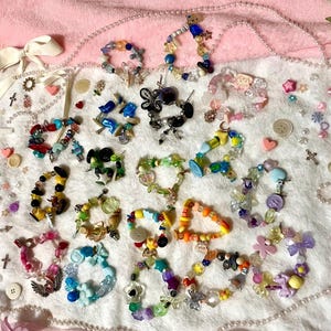 May include: A collection of colorful beaded bracelets with various charms, buttons, and beads. The bracelets are arranged on a soft pink surface, showcasing a variety of designs and colors, including blue, yellow, green, and purple.