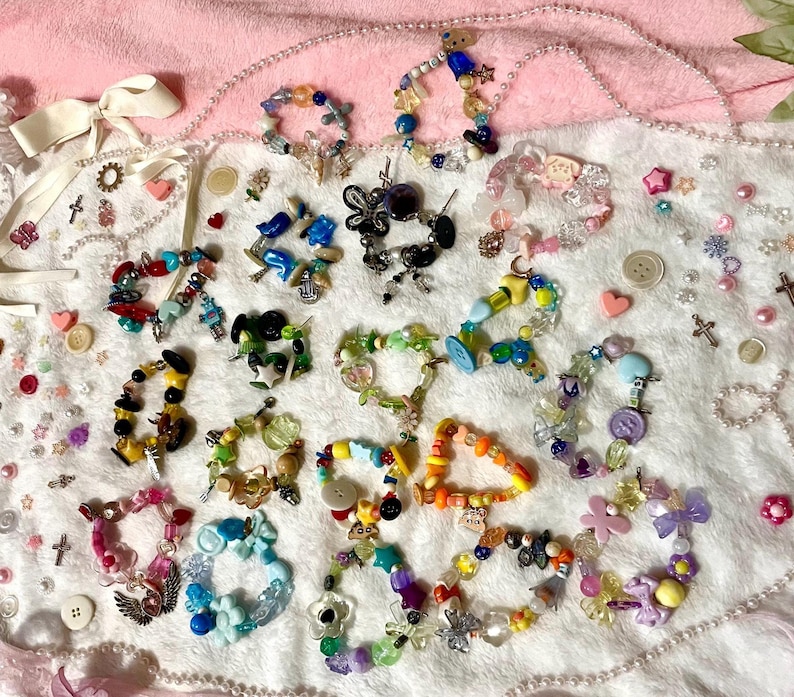 May include: A collection of colorful beaded bracelets with various charms, buttons, and beads. The bracelets are arranged on a soft pink surface, showcasing a variety of designs and colors, including blue, yellow, green, and purple.
