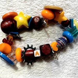 May include: A colorful beaded bracelet with a variety of shapes and colors. The bracelet includes wooden beads, a yellow star, orange and blue beads, and a bead with the letter "A".