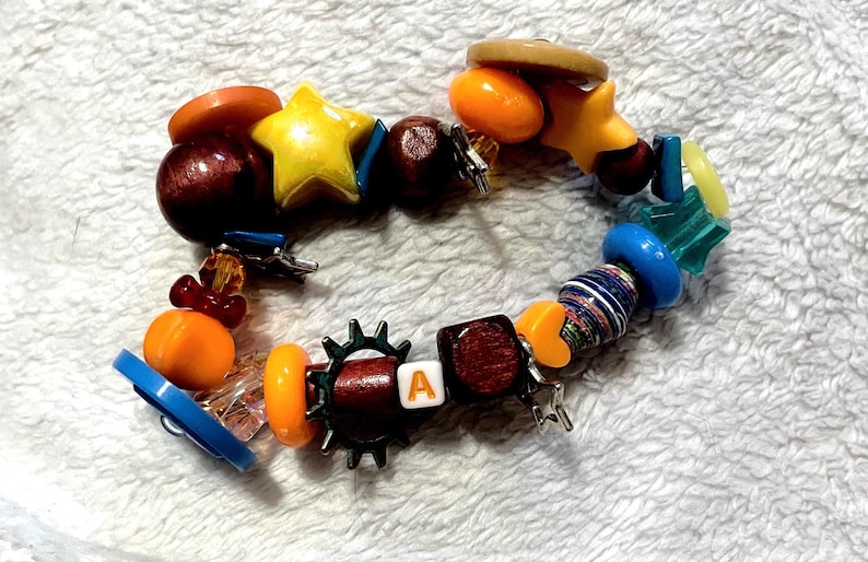 May include: A colorful beaded bracelet with a variety of shapes and colors. The bracelet includes wooden beads, a yellow star, orange and blue beads, and a bead with the letter "A".