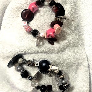 May include: Two handmade bracelets with a variety of beads and charms. One bracelet features pink star-shaped beads, black buttons, and a silver charm. The other has black and silver beads, a large black bead, and a white die.