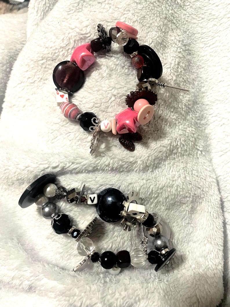 May include: Two handmade bracelets with a variety of beads and charms. One bracelet features pink star-shaped beads, black buttons, and a silver charm. The other has black and silver beads, a large black bead, and a white die.