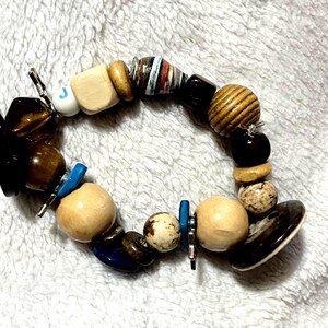 May include: A beaded bracelet featuring a variety of beads in different shapes, sizes, and materials. The bracelet includes wooden beads, dark brown beads, and beads with a marbled appearance. Accents include blue and silver metal elements.