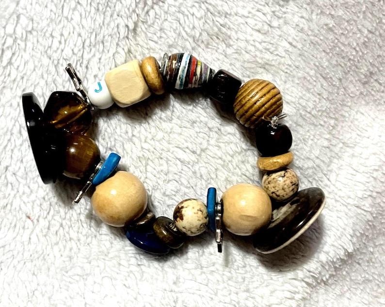 May include: A beaded bracelet featuring a variety of beads in different shapes, sizes, and materials. The bracelet includes wooden beads, dark brown beads, and beads with a marbled appearance. Accents include blue and silver metal elements.
