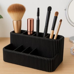 Könnte beinhalten: Ein schwarzer, geriffelter Make-up-Organizer enthält verschiedene Pinsel, einen Lippenstift und einen Mascara-Stab. Ein großer, flauschiger Pinsel und ein kleiner Spiegel sind ebenfalls sichtbar. Der Organizer steht auf einer hellen Holzoberfläche, mit einer Topfpflanze im Hintergrund.