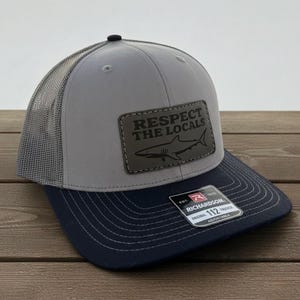 May include: Gray and navy blue trucker hat with a mesh back. The hat features a rectangular patch with the text "RESPECT THE LOCALS" and a shark graphic. The brim is navy blue, and the hat is adjustable.