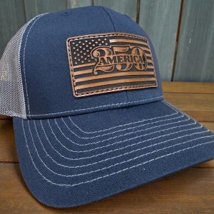 May include: Navy blue trucker hat with a white mesh back and a curved brim. The front features a brown leather patch with an American flag design and the text "250 AMERICA" in a vintage style.