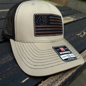 May include: Tan and black trucker hat with a brown leather patch featuring a stylized American flag with the numbers "45" and "47". The hat has a mesh back and a curved brim. The tag reads "Richardson 112".