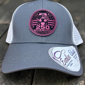 May include: Gray trucker hat with a white mesh back and a pink patch featuring a dog wearing sunglasses, stars, and the text "250 YEARS OF FREEDOM 1776-2026." The hat also has a tag with the text "Evolve Elude."