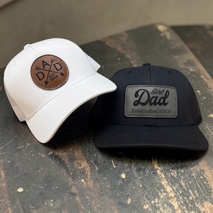 May include: A white baseball cap with a round brown leather patch featuring the text DAD with crossed arrows and est. 2018, and a black baseball cap with a rectangular black leather patch featuring the text Girl Dad Established 2006.
