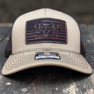May include: Tan and brown trucker hat with a leather patch featuring the American flag and the text "250 AMERICA 1776-2026". The hat has a mesh back and a curved brim. The Richardson brand tag is visible.