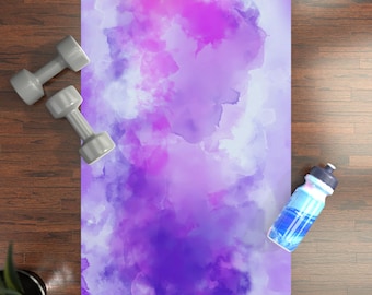 May include: A yoga mat with a watercolor design in shades of purple and white. Two gray dumbbells and a blue water bottle are on the wooden floor next to the mat. The mat is designed for fitness and exercise.