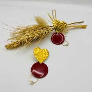 Könnte beinhalten: Ein Paar goldene herzförmige Ohrringe mit strukturierter Oberfläche, kombiniert mit einem tiefroten kreisförmigen Anhänger. Die Ohrringe sind durch eine feine Goldkette mit einem kleinen, funkelnden Akzent verbunden. Auf weißem Hintergrund.