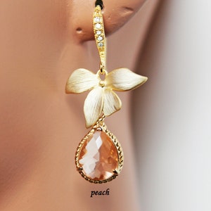 May include: Gold-toned drop earrings featuring a peach-colored teardrop-shaped gemstone. The earrings have a floral design with a textured finish and are accented with small, clear crystals. The word "peach" is written on the image.