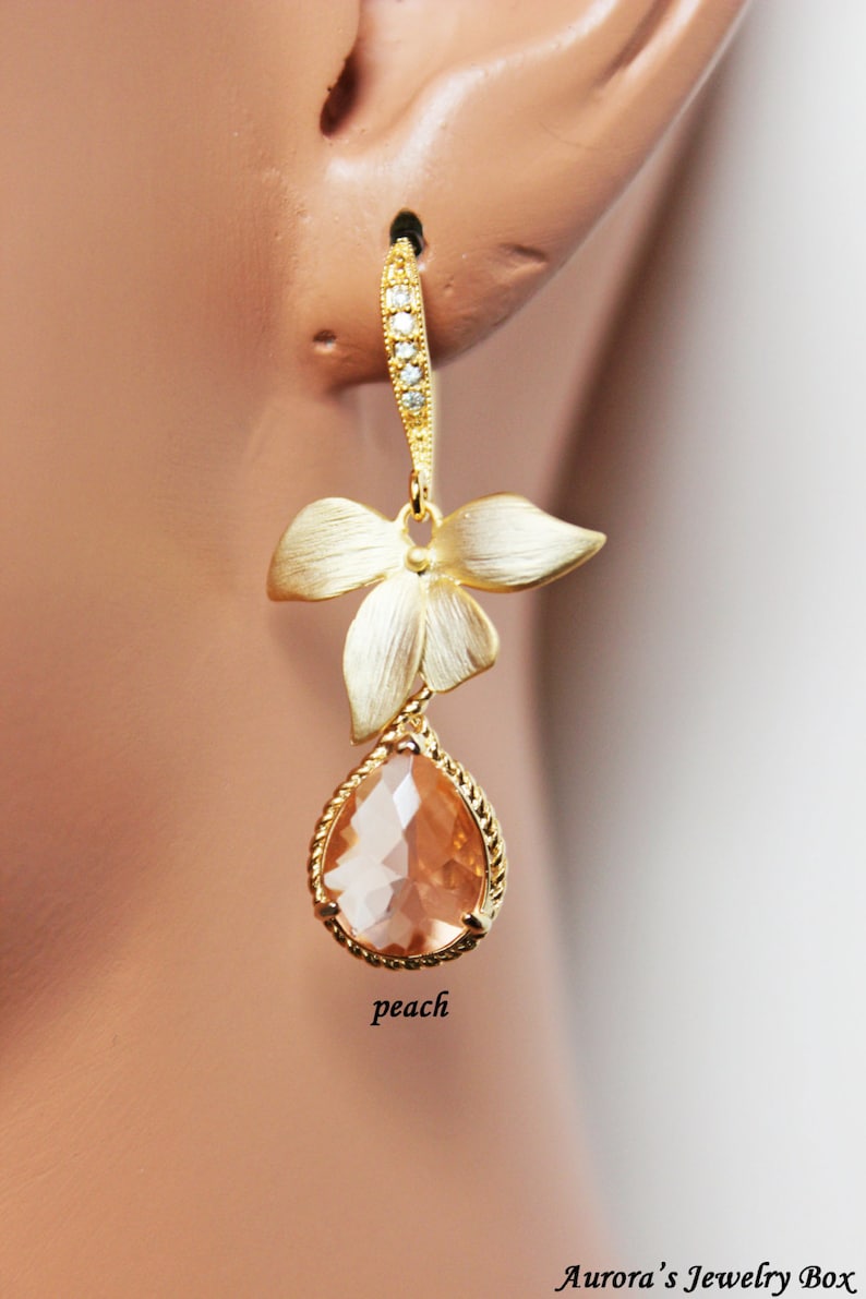 May include: Gold-toned drop earrings featuring a peach-colored teardrop-shaped gemstone. The earrings have a floral design with a textured finish and are accented with small, clear crystals. The word "peach" is written on the image.