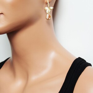 May include: Gold-toned drop earrings featuring a floral design and a teardrop-shaped, amber-colored stone. The earrings are worn on a mannequin, showcasing the jewelry's design and color against a neutral background. The earrings are a stylish accessory.