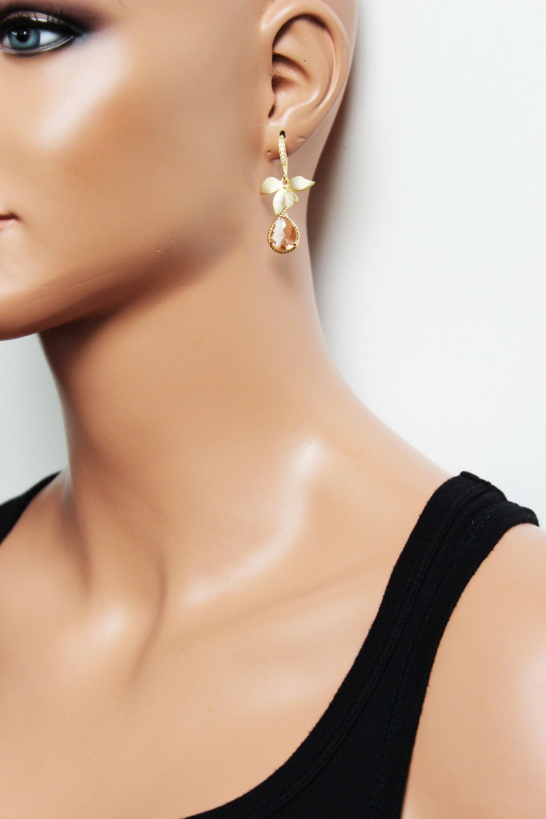 May include: Gold-toned drop earrings featuring a floral design and a teardrop-shaped, amber-colored stone. The earrings are worn on a mannequin, showcasing the jewelry's design and color against a neutral background. The earrings are a stylish accessory.