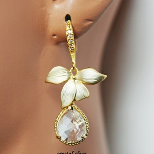 May include: A gold-toned earring with a floral design and a clear, teardrop-shaped crystal. The earring features a textured finish on the flower petals and a beaded edge around the crystal. The text "crystal clear" is visible.