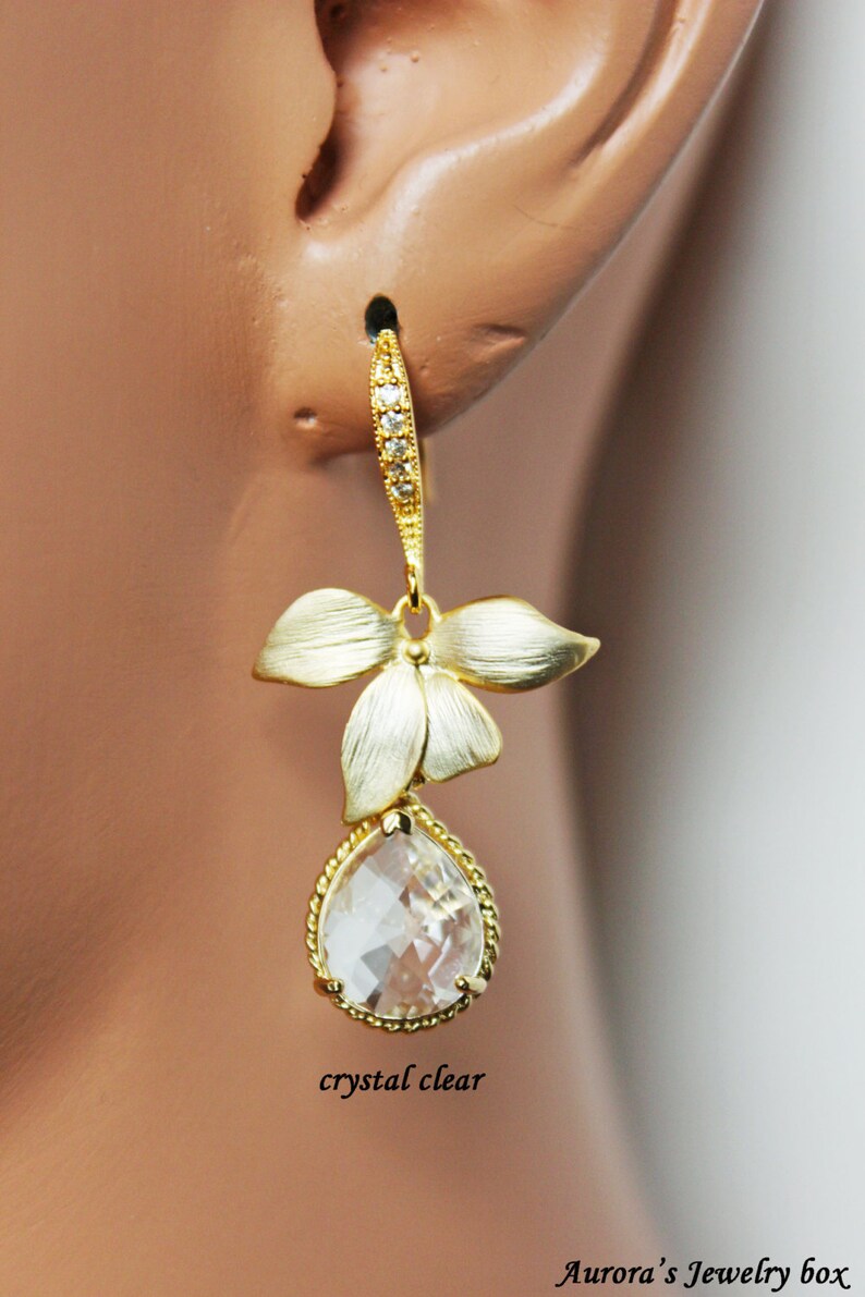 May include: A gold-toned earring with a floral design and a clear, teardrop-shaped crystal. The earring features a textured finish on the flower petals and a beaded edge around the crystal. The text "crystal clear" is visible.