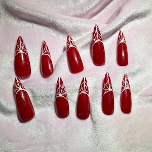 May include: A set of ten red, pointed false nails with a white spiderweb design near the tips. The nails are arranged on a pink, textured surface, showcasing a Halloween or gothic theme.
