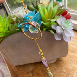 May include: A handcrafted gold-colored metal suncatcher with a clear crystal orb and amethyst and green gemstone accents. The suncatcher is suspended from a delicate chain and is displayed in front of a wooden planter filled with colorful artificial flowers.